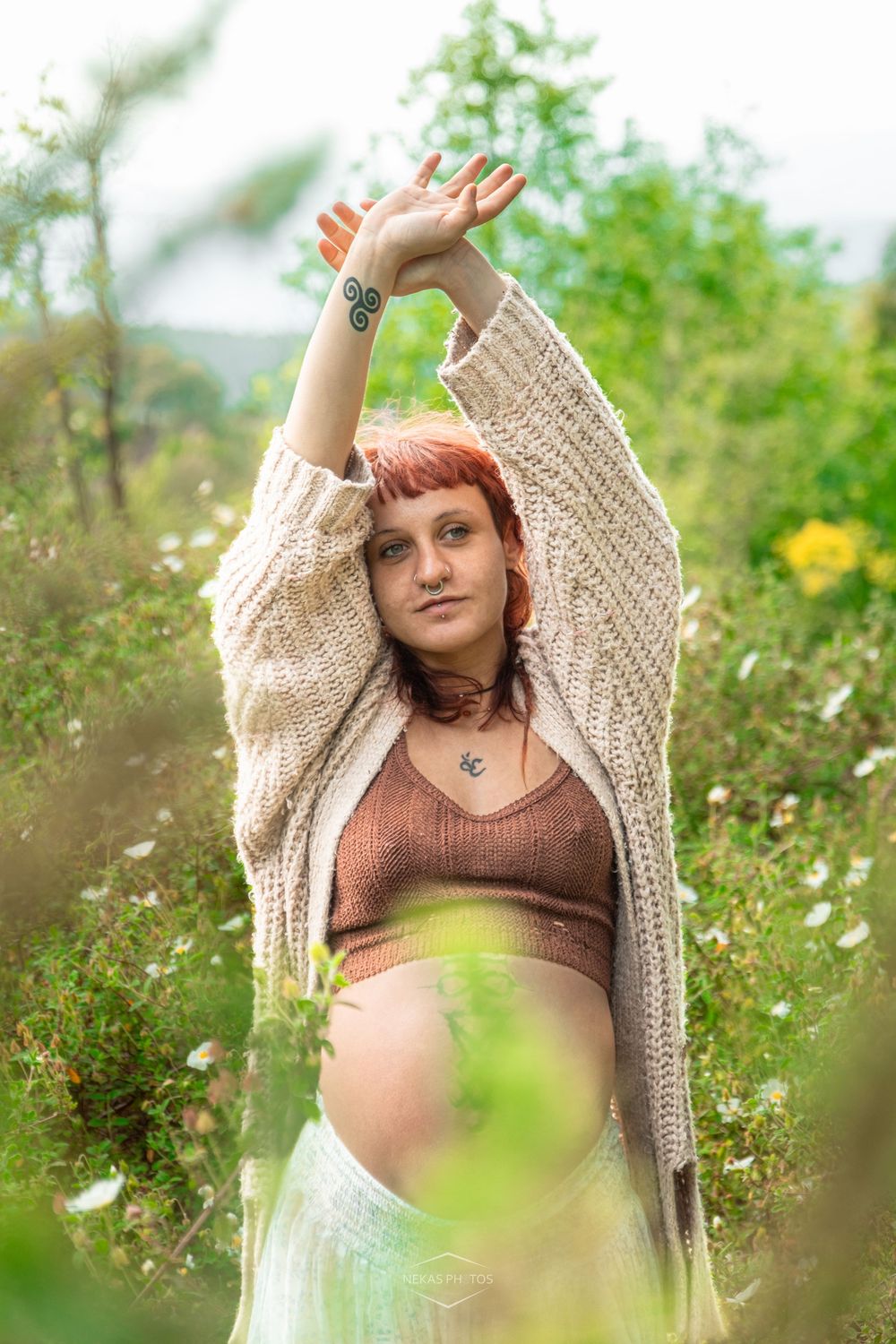 Pregnant woman with red hair poses outdoors in a floral field, wearing a knit cardigan and crop top, arms raised gracefully.