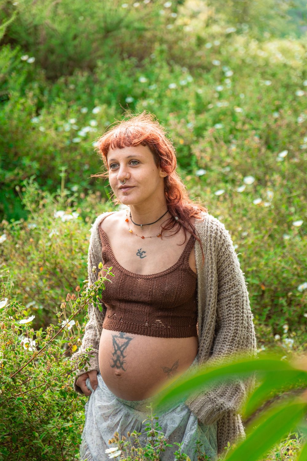 Pregnant woman with red hair and tattoos in a brown crop top and cardigan standing among wildflowers outdoors.