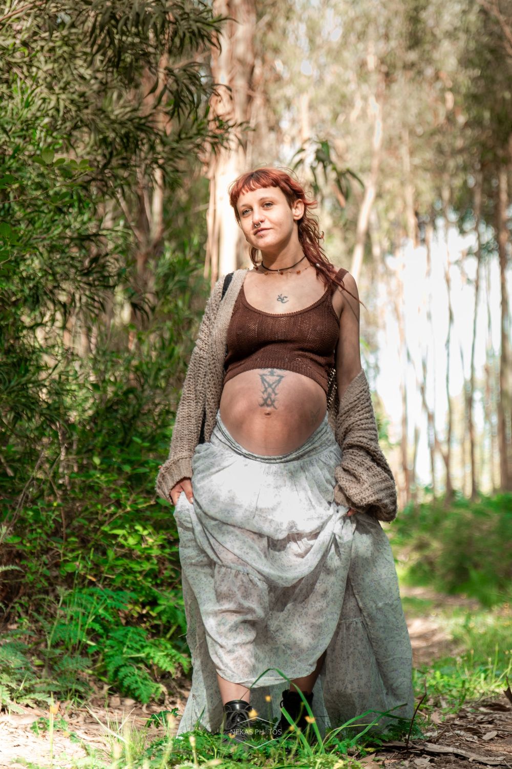 Pregnant woman with red hair stands in a forest wearing a brown crop top, gray skirt, and cardigan, showing her bare belly.