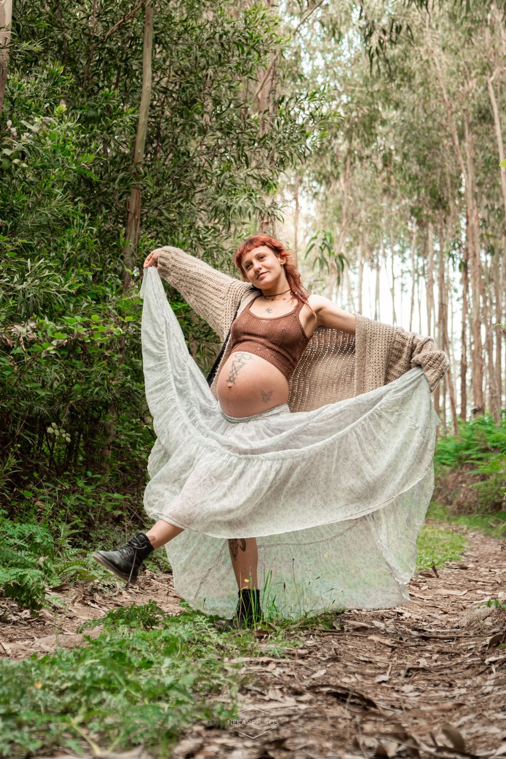 Pregnant redhead woman in white lace skirt poses gracefully on a forest path surrounded by tall trees.
