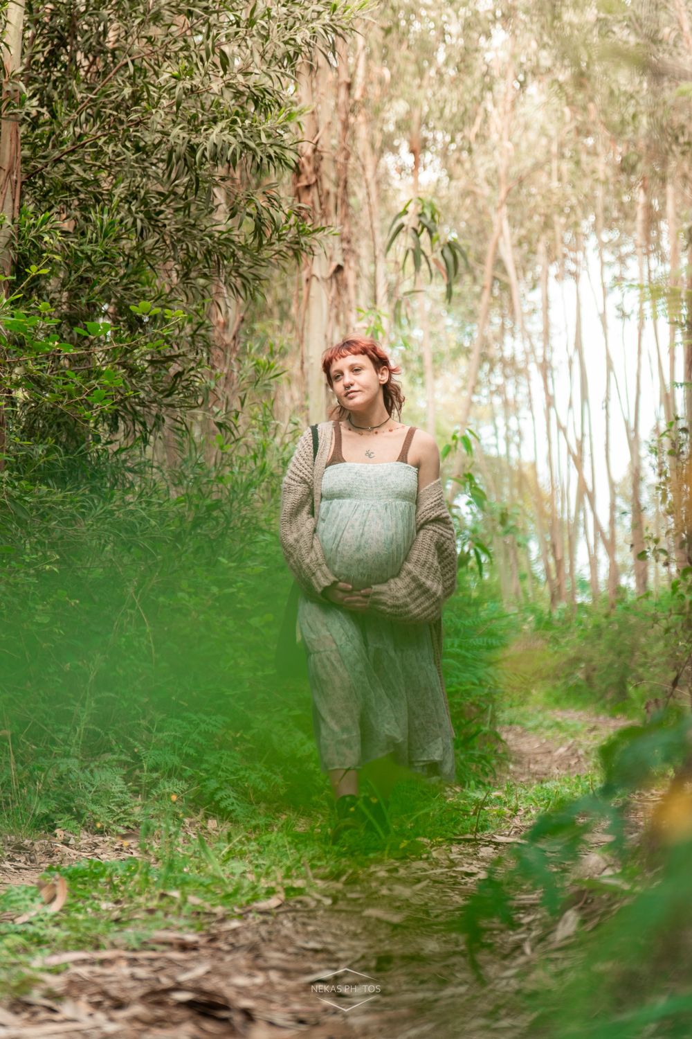 Pregnant woman in sage green dress and cardigan standing on a forest path surrounded by lush green trees.