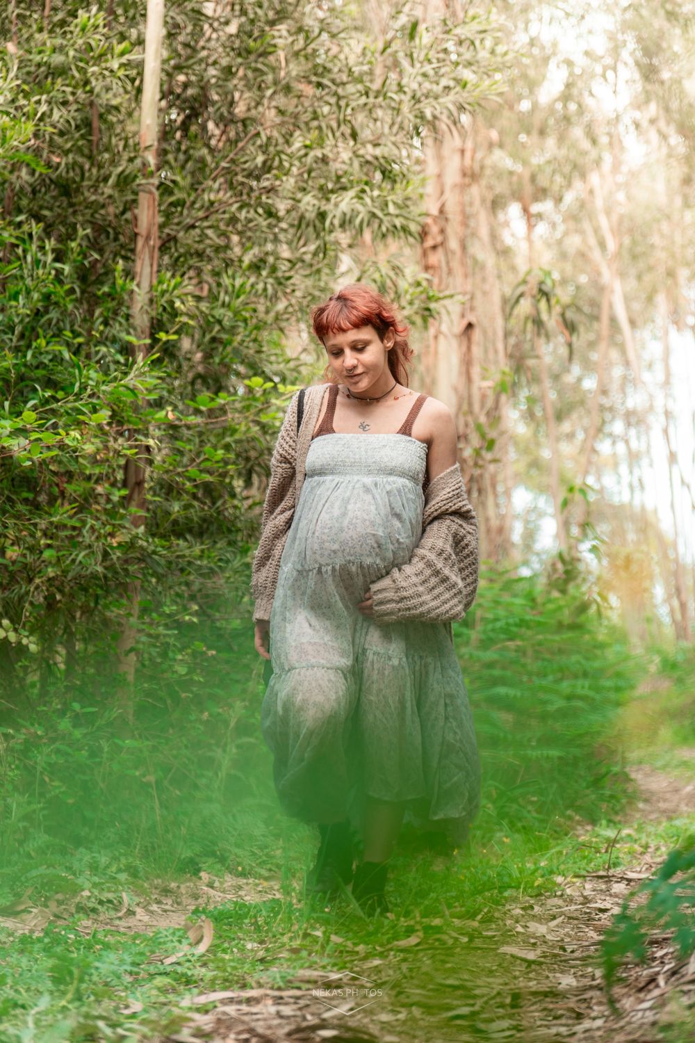 Pregnant woman in green tulle dress and knit cardigan walks through a lush forest path with green smoke effect.