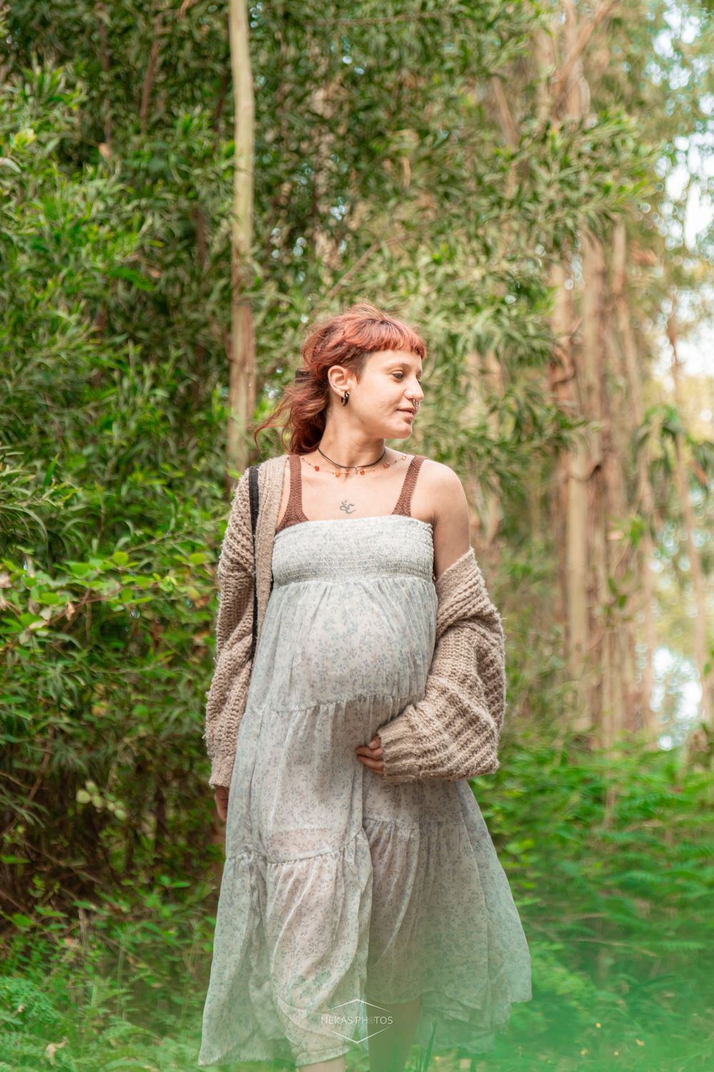 Pregnant woman in gray dress and brown cardigan standing in lush green forest path, cradling her baby bump.
