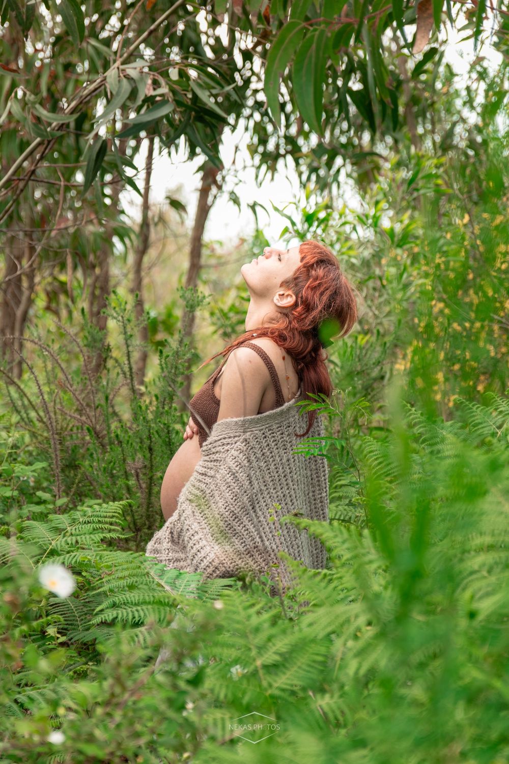 Red-haired woman sitting among lush green ferns and trees, head tilted back with eyes closed in nature.