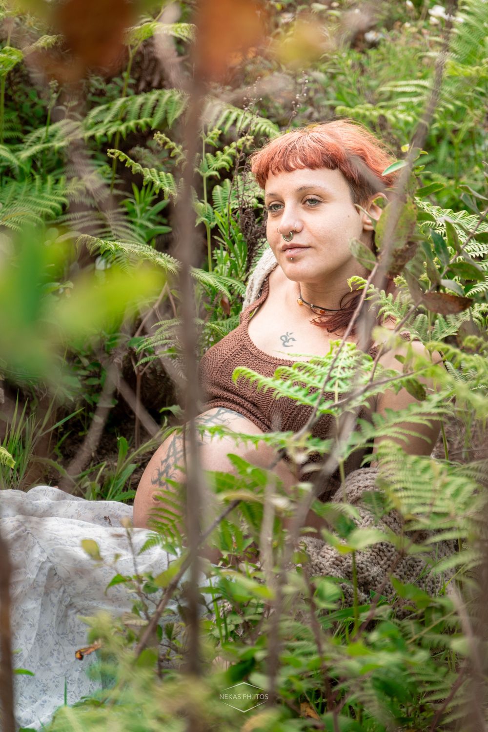 Young woman with red hair and elf ears sits among lush green ferns in a forest, wearing a brown top.