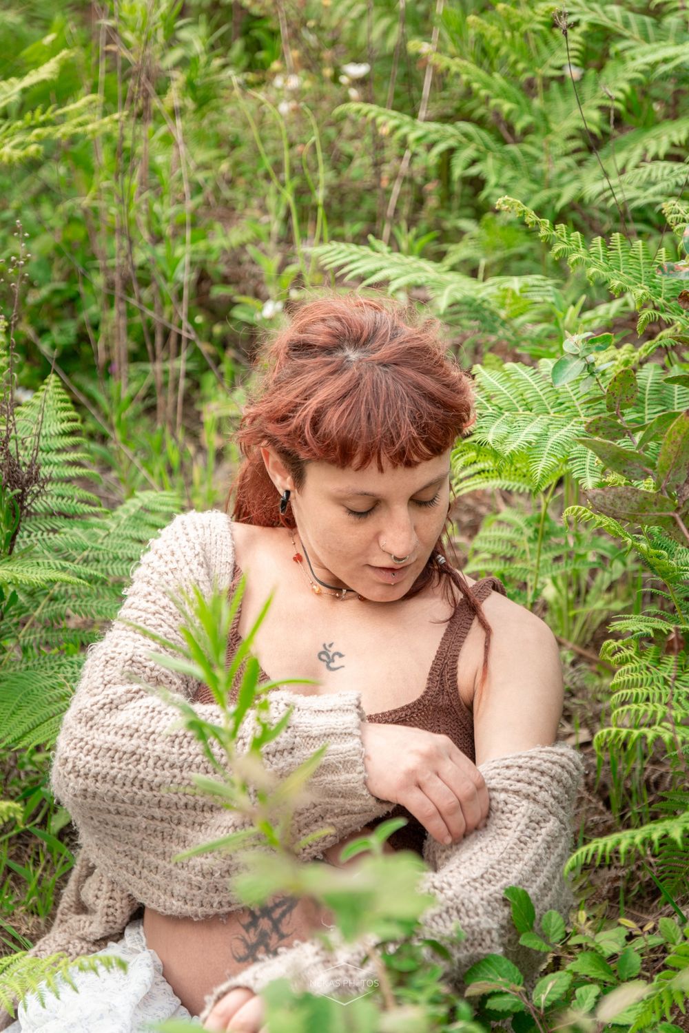 Red-haired woman in knit cardigan sitting among lush green ferns and plants in a forest setting.