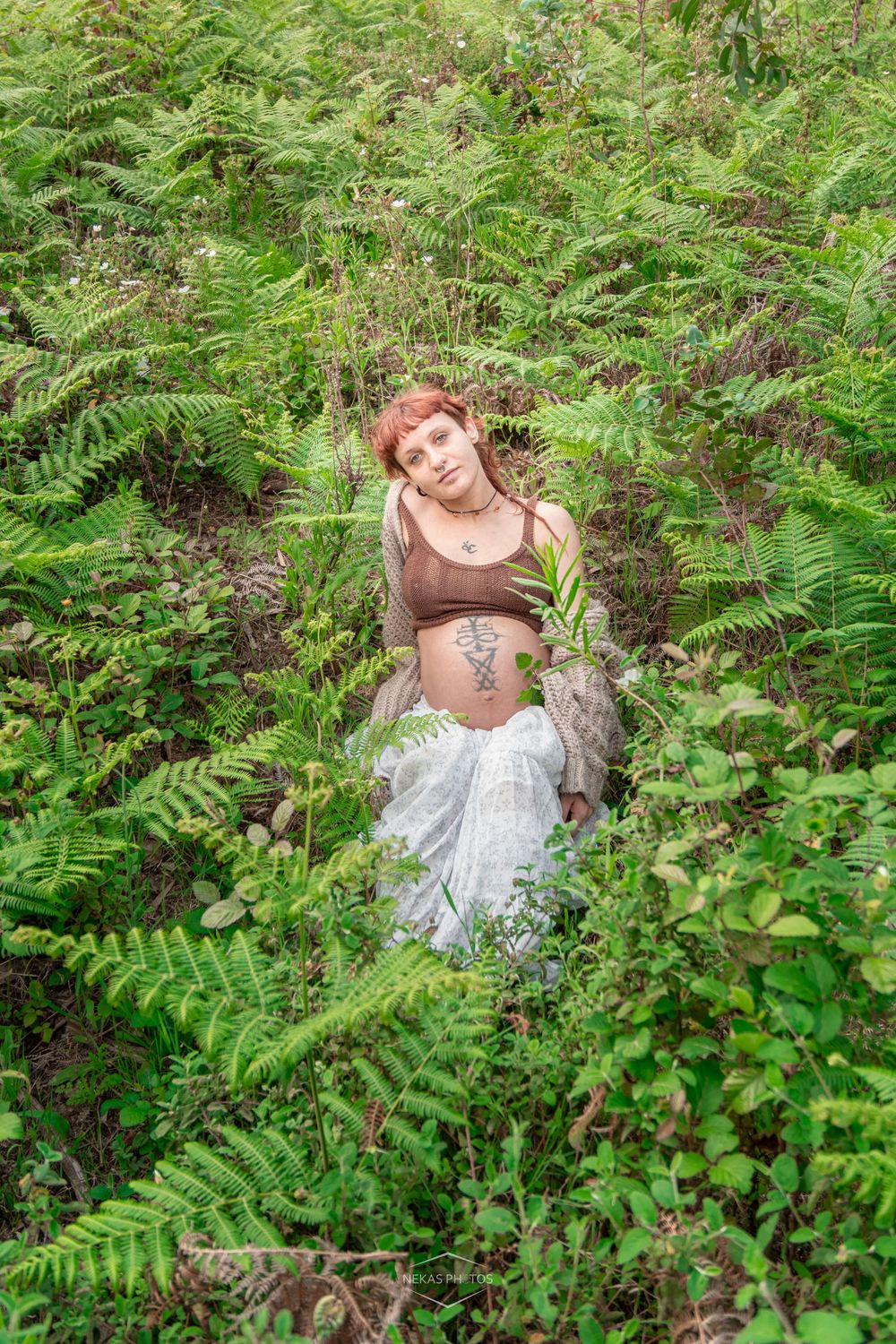 Pregnant woman with red hair sitting among lush green ferns outdoors, wearing a brown crop top and white skirt.