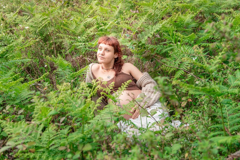 A young redhead woman sits peacefully among lush green ferns in a forest, gazing upward in a serene outdoor setting.