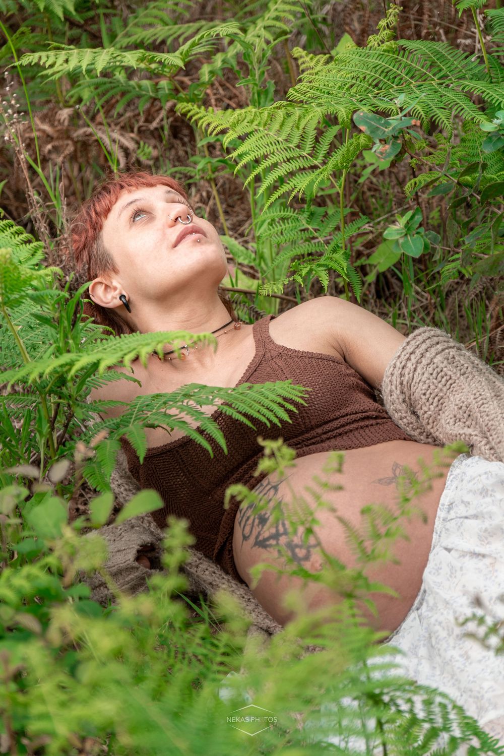 Woman with short red hair lying among green ferns and foliage, wearing a brown top, looking upward peacefully.