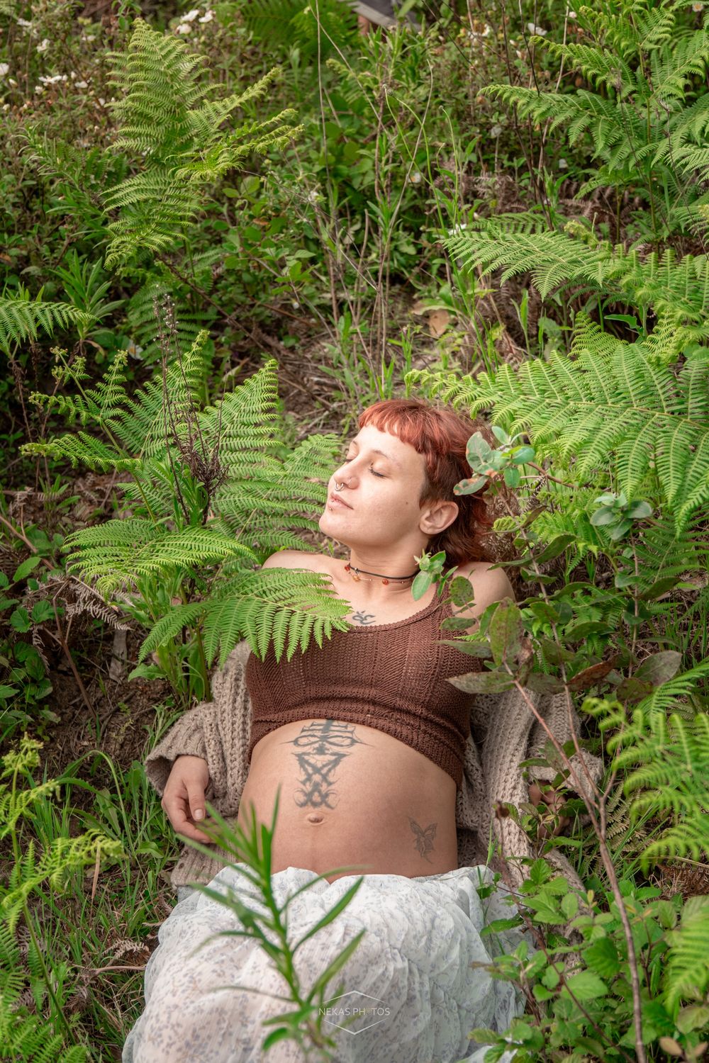 Pregnant woman with tattoos lying peacefully among lush green ferns and plants outdoors.