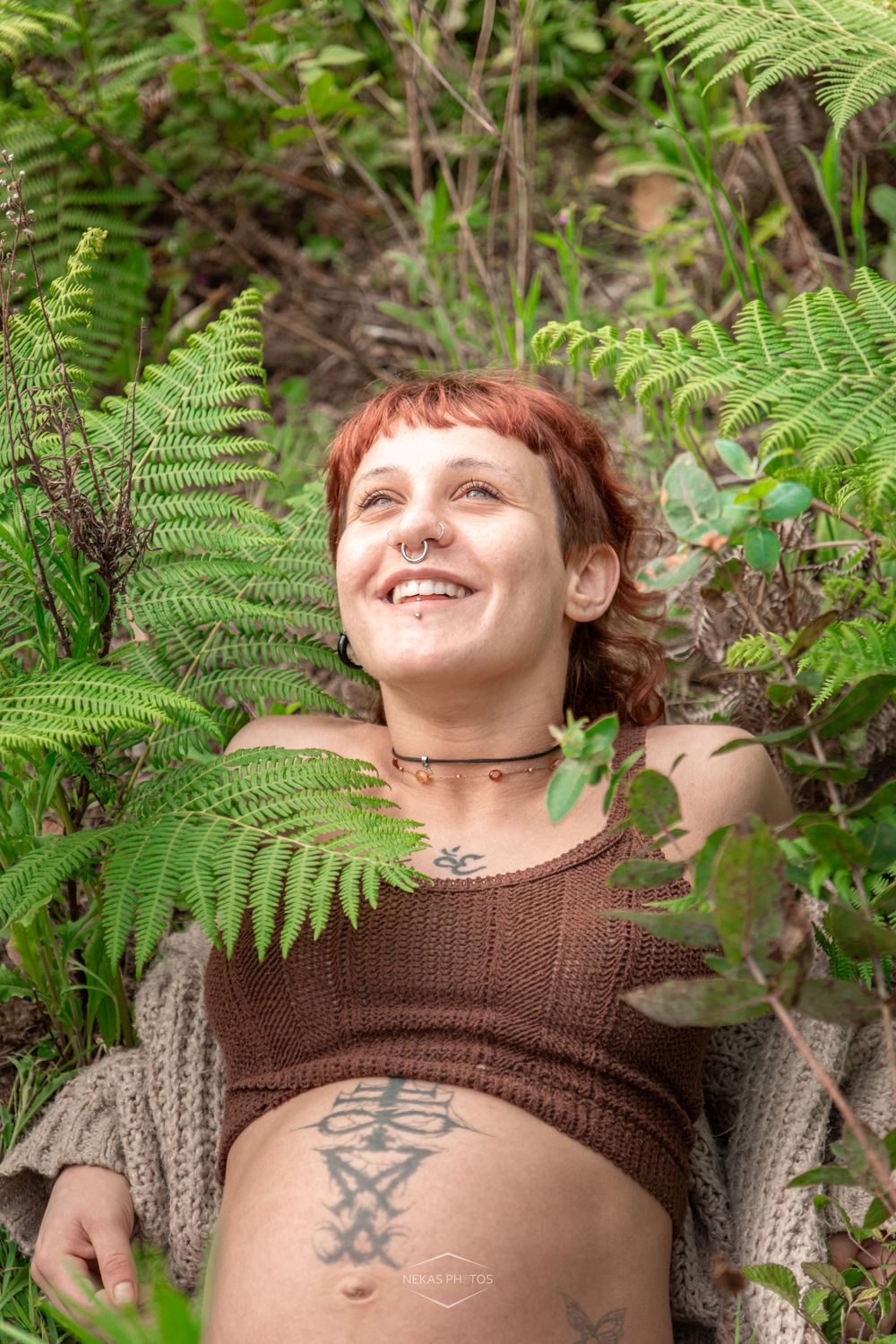 Smiling redhead woman with tattoos lying among lush green ferns, wearing a brown crop top and cardigan outdoors.