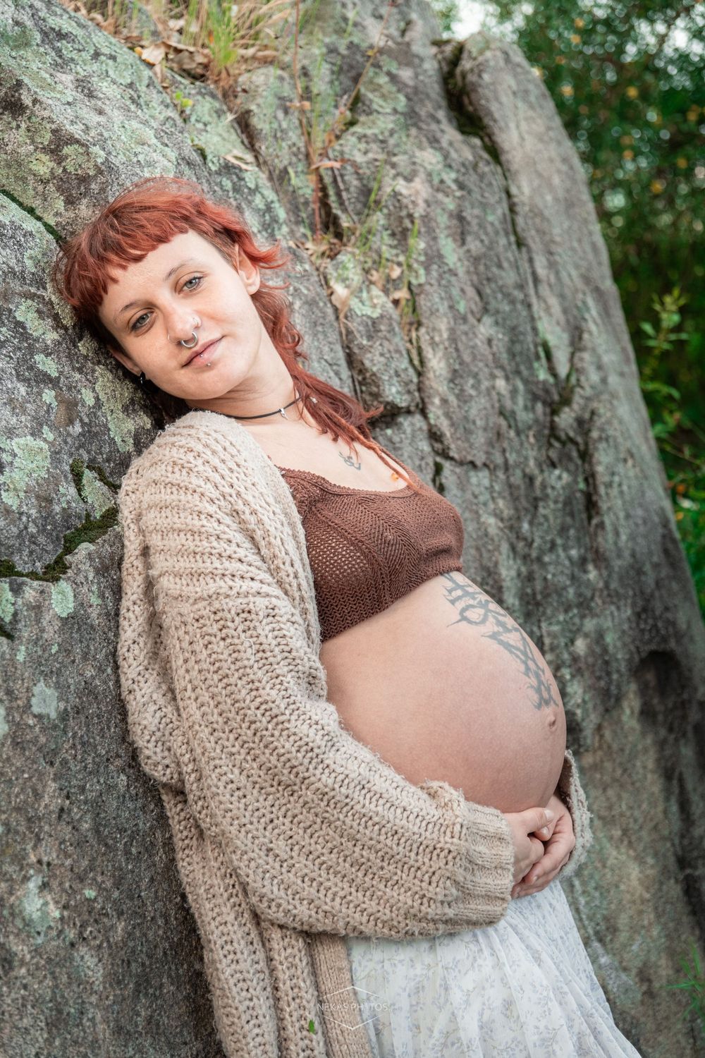 Pregnant redhead woman leaning against mossy rocks outdoors, cradling her baby bump in a cozy knit cardigan.