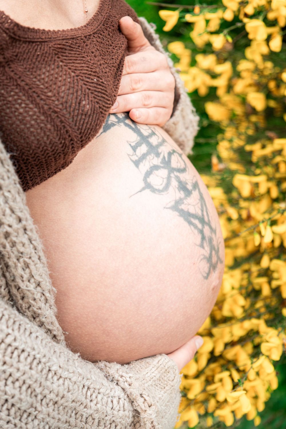 Pregnant woman outdoors cradling bare baby bump with tattoo, wearing brown crop top and beige cardigan near yellow flowers.