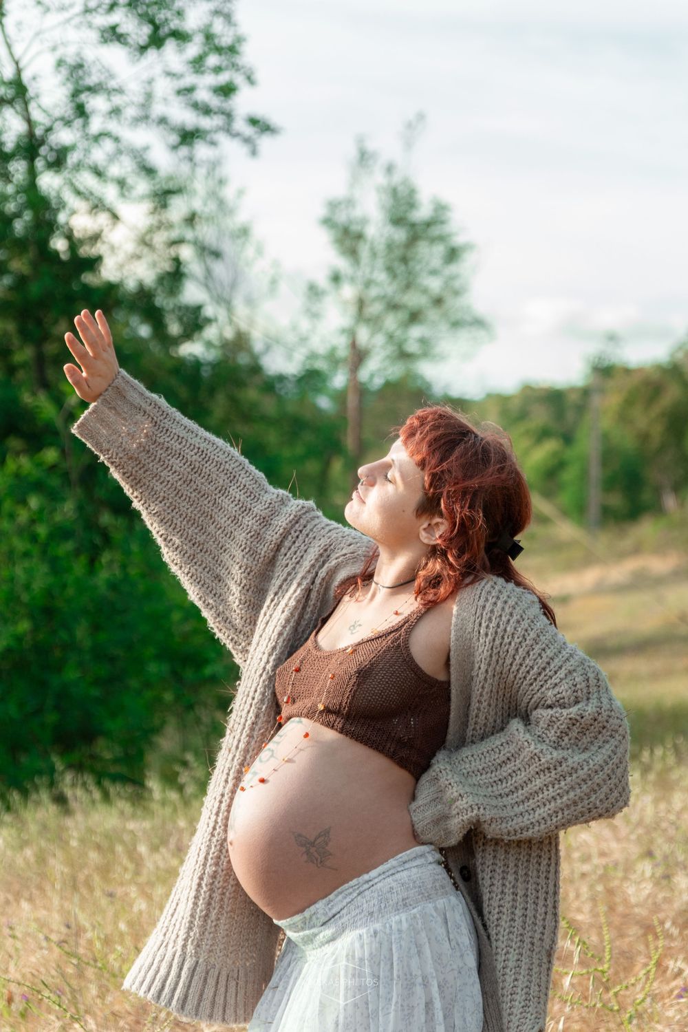 Pregnant woman with red hair in knit cardigan and crop top poses outdoors in field, arm raised toward sky.