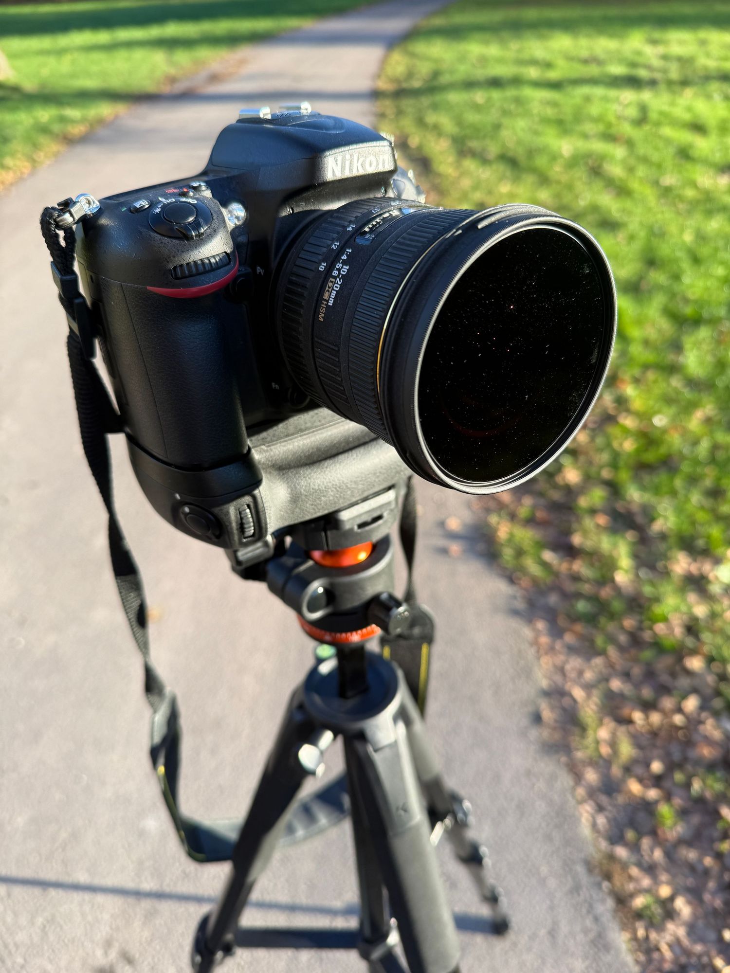 Nikon DSLR camera with large lens mounted on tripod on a paved path outdoors.