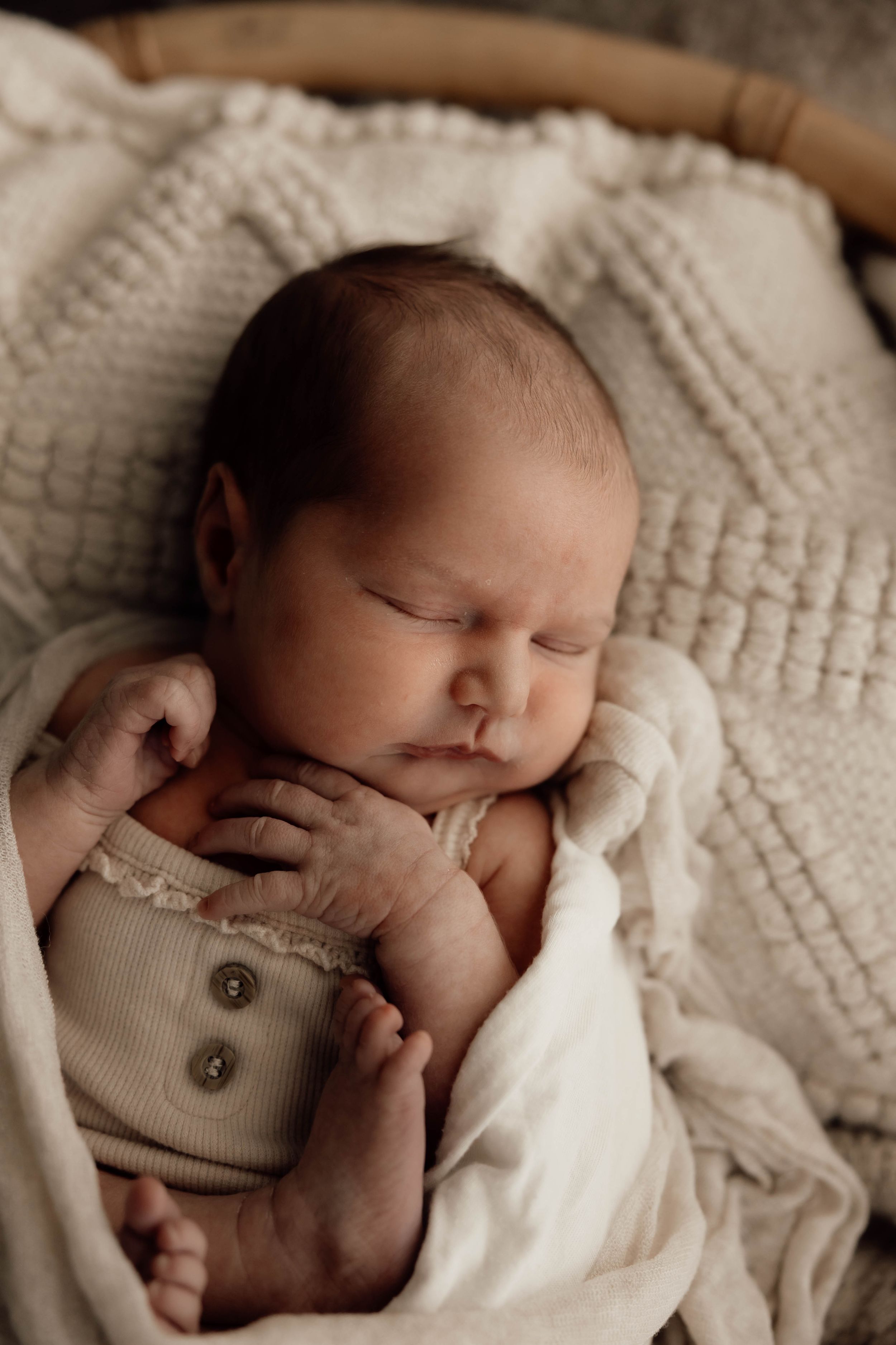 Sleeping newborn baby wrapped in cream muslin swaddle wearing sage green ribbed onesie in wicker basket.