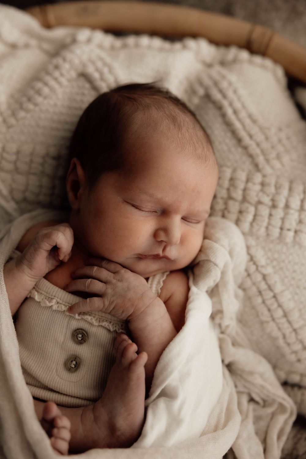 Sleeping newborn baby wrapped in cream muslin swaddle wearing sage green ribbed onesie in wicker basket.
