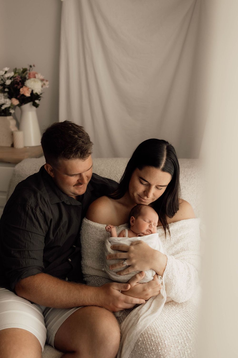 New parents sitting on a couch lovingly gazing at their sleeping newborn wrapped in white.