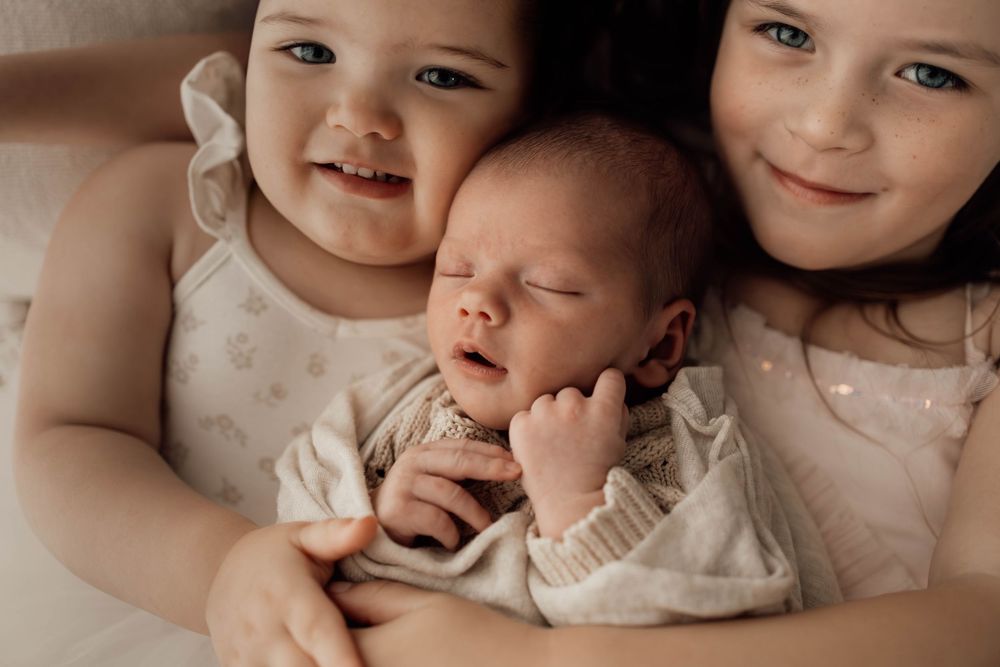 Two young sisters gently cradle their sleeping newborn sibling in a tender family portrait.