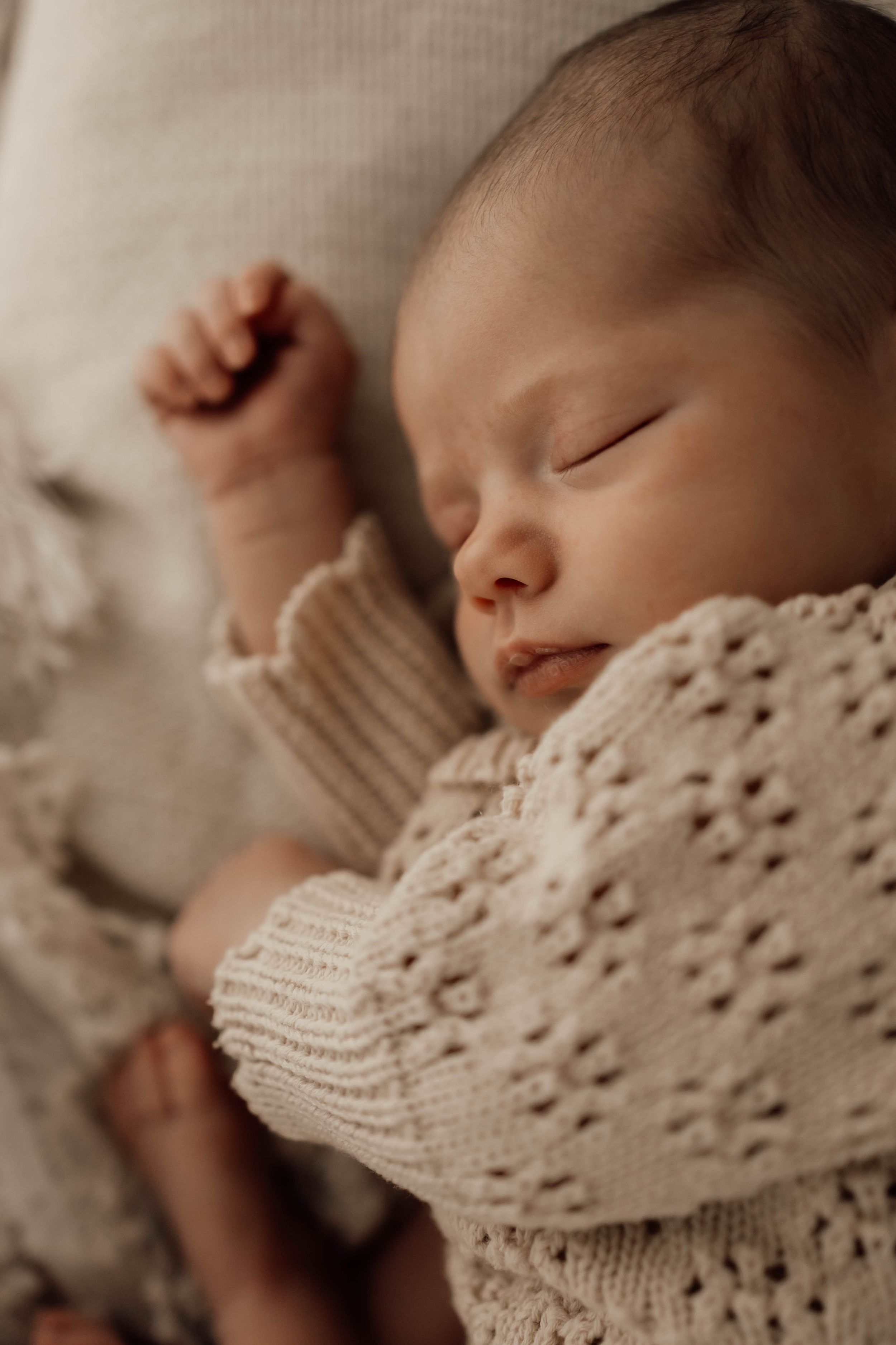 Close-up of a peacefully sleeping newborn in a beige knit romper with tiny fist raised.