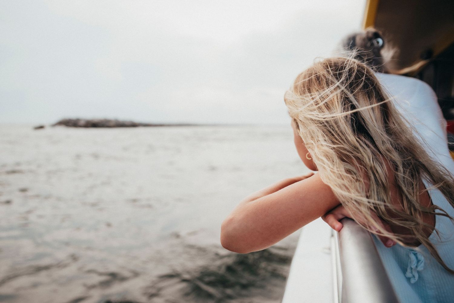 Young girl with blonde hair leaning over a boat railing, gazing at the ocean and rocky shore.