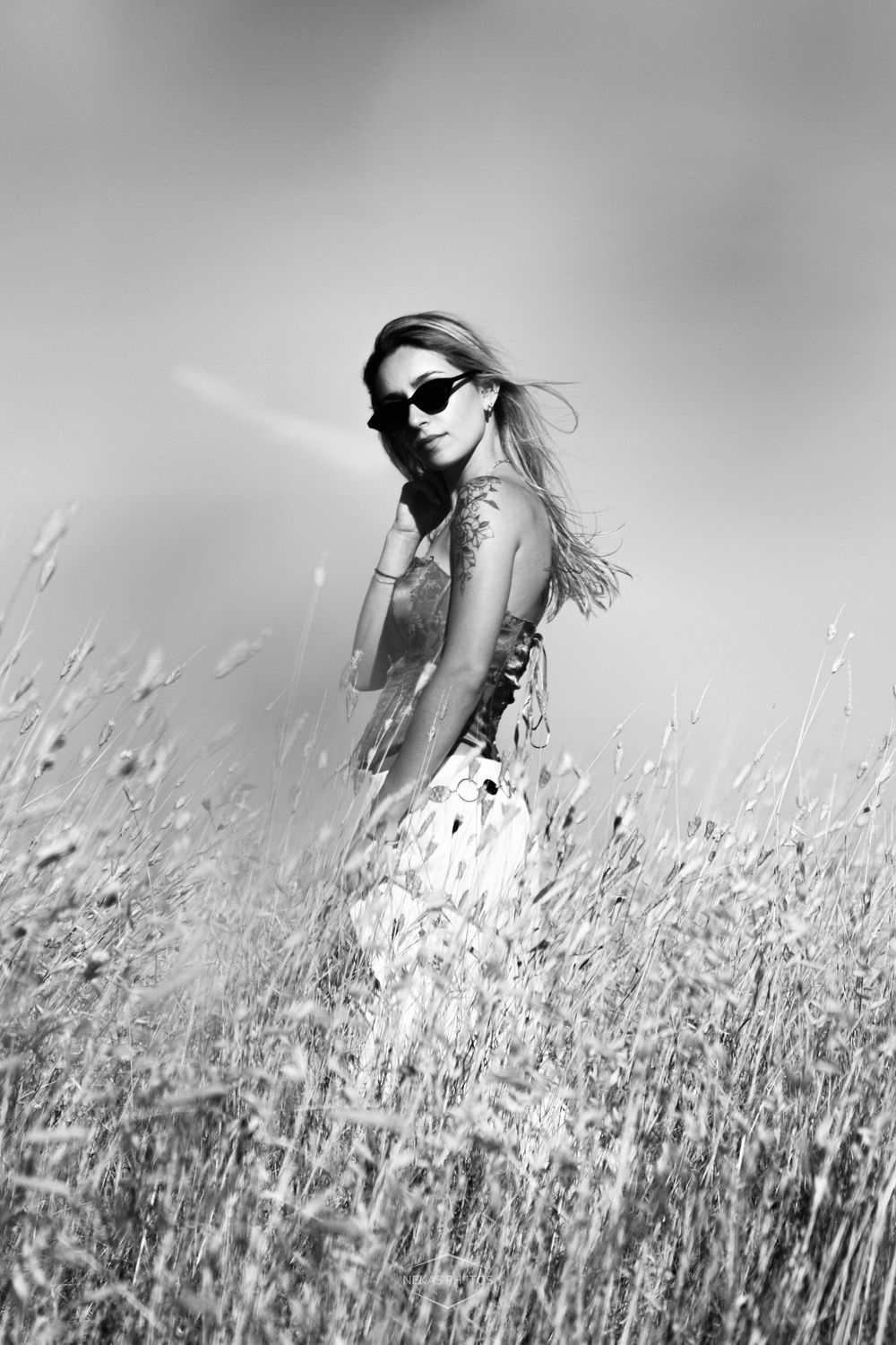 Black and white photo of a woman in sunglasses and floral dress standing in tall grass field with wind-blown hair.