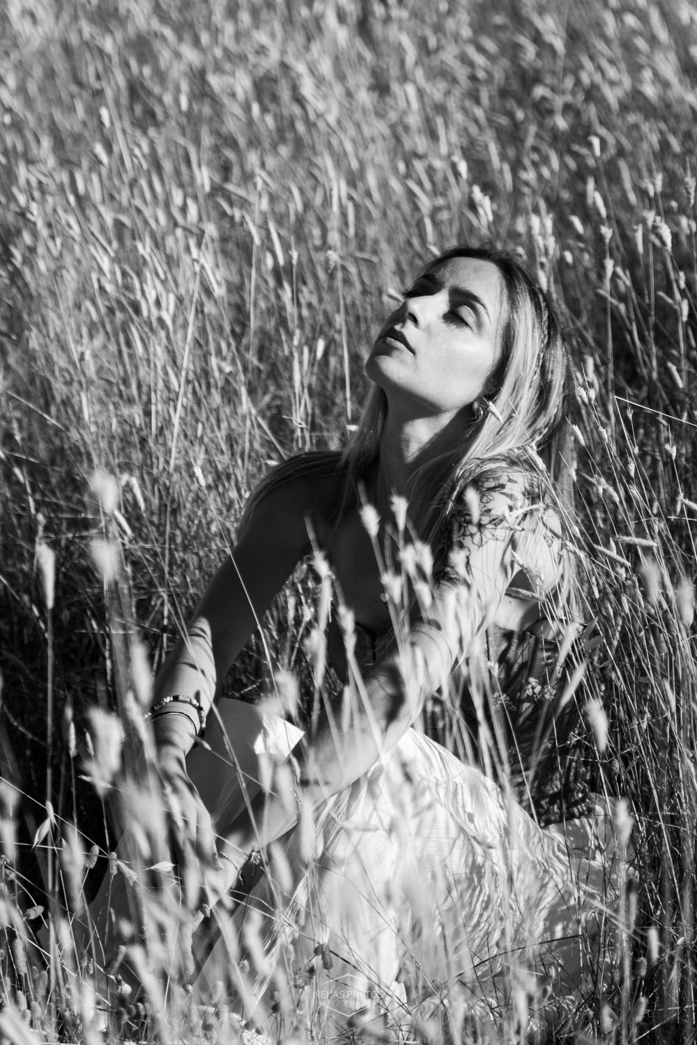 Black and white photo of a woman sitting in tall grass field, looking up with long hair, surrounded by wild wheat.