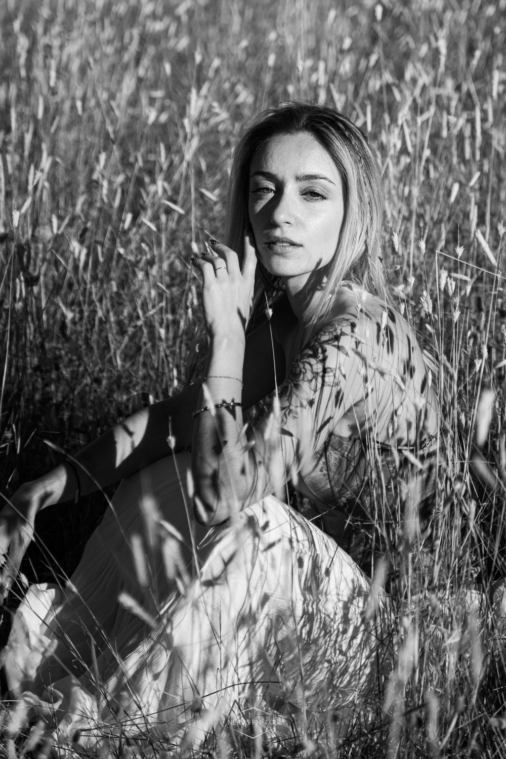 Black and white portrait of a young woman sitting in tall grass field, hand near face, wearing patterned dress.