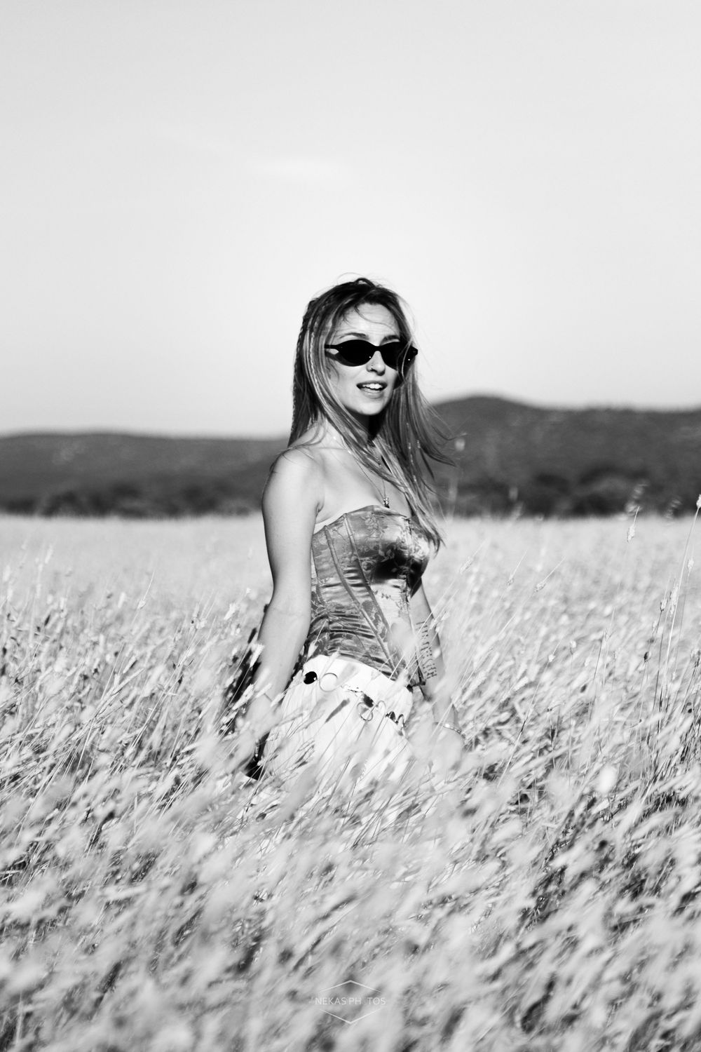 Black and white photo of a woman in sunglasses and strapless top standing in a golden wheat field.