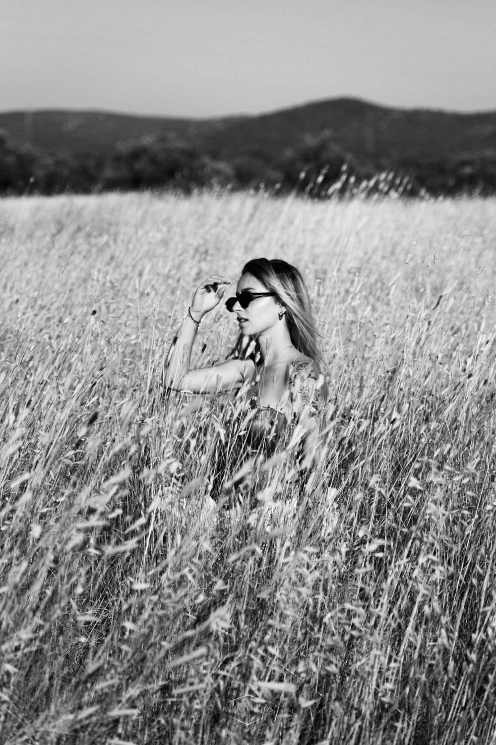 Black and white photo of a woman in sunglasses holding a camera in a golden wheat field with hills in the background.