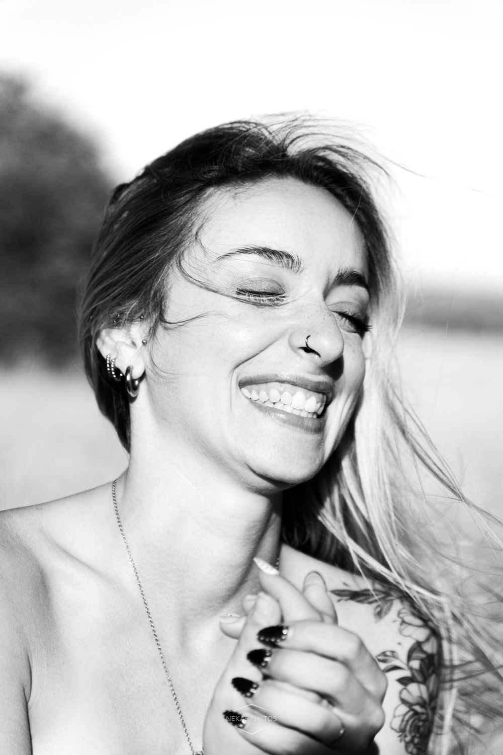 Black and white portrait of a laughing woman with tattoos, nose ring, and windswept hair outdoors.
