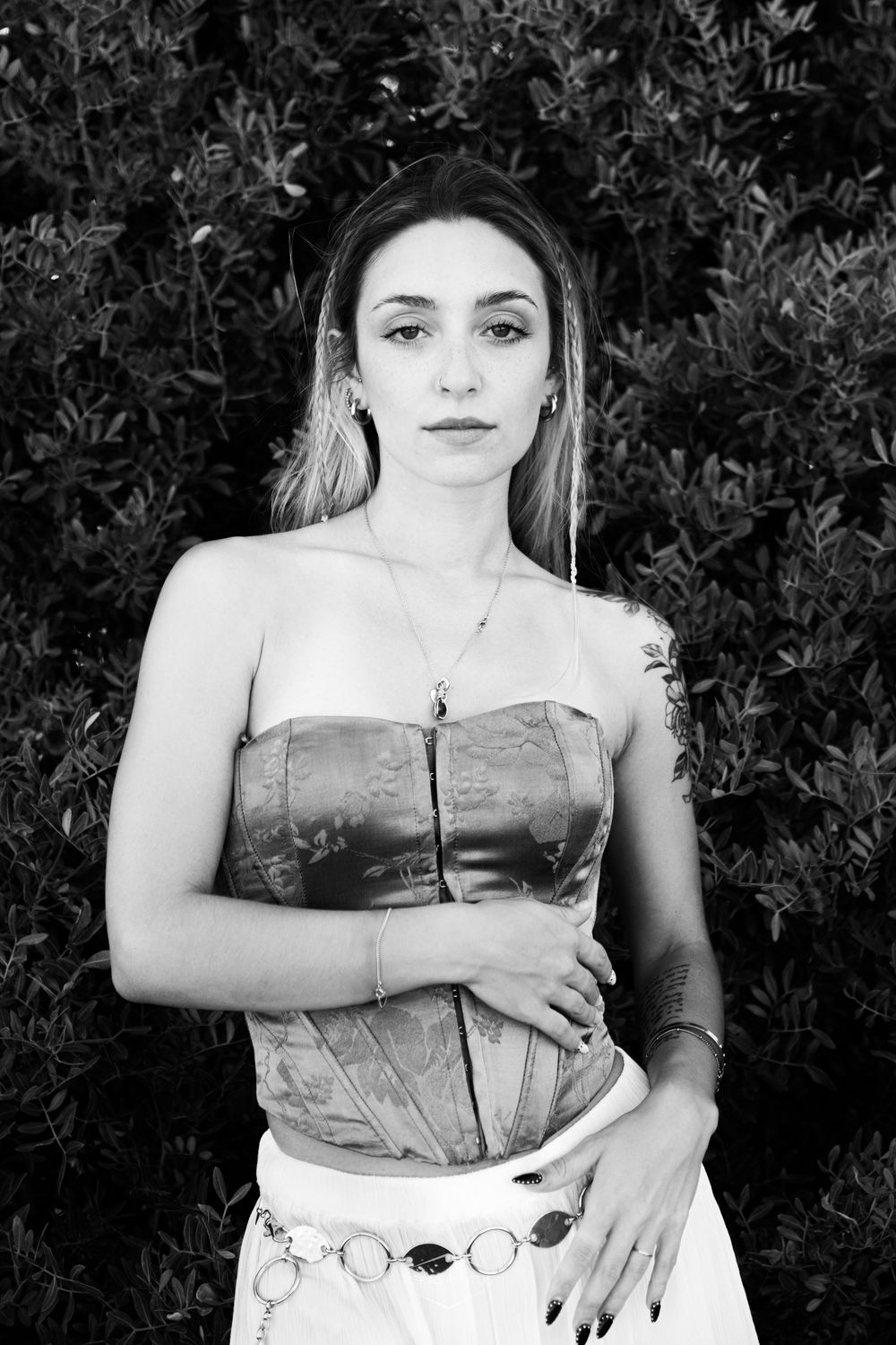 Black and white portrait of a young woman in a strapless corset top with jewelry, posing before lush green bushes.