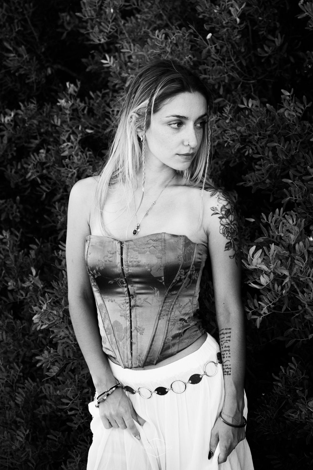 Black and white portrait of a young woman with tattoos wearing a corset top and white skirt, posing outdoors near lush green bushes.