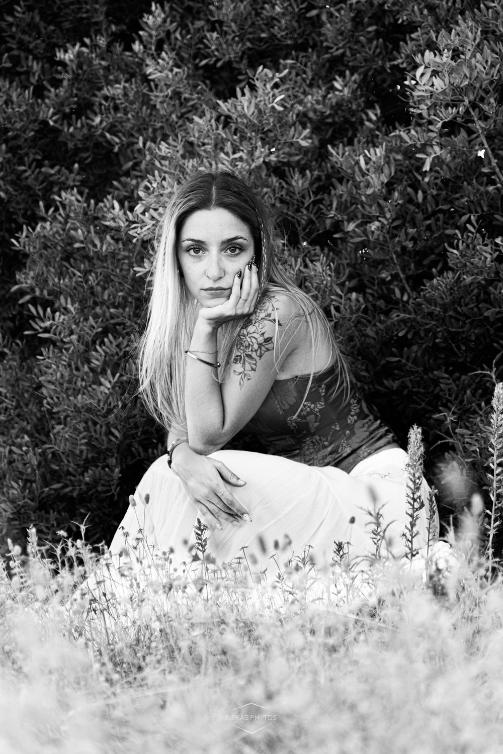 Black and white portrait of a young woman with long hair sitting in a field, resting her chin on her hand.