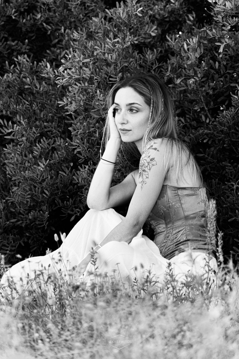 Black and white portrait of a young woman with a tattoo sitting in a field of wildflowers near lush green bushes.