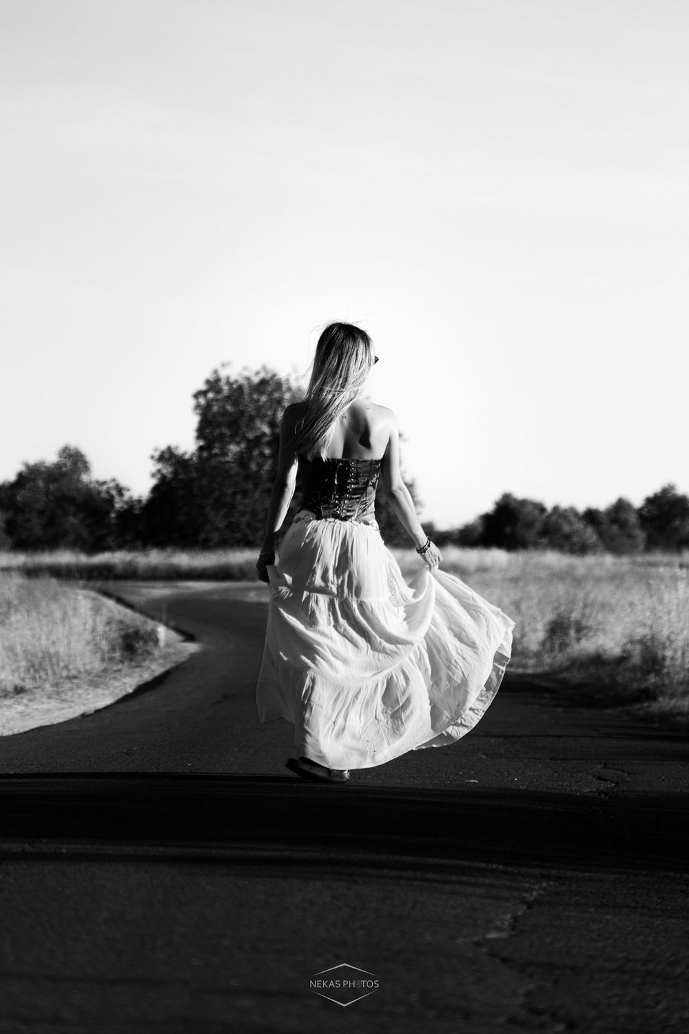 Black and white photo of a woman in a corset and flowing skirt walking away on a rural road.