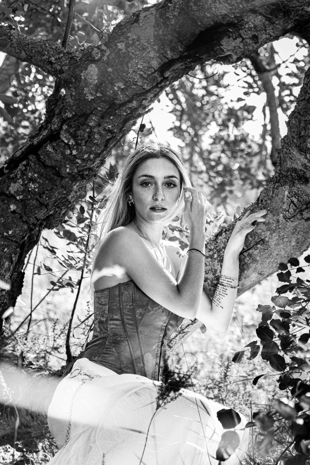 Black and white portrait of a young woman in a corset posing gracefully among tree branches in a forest.