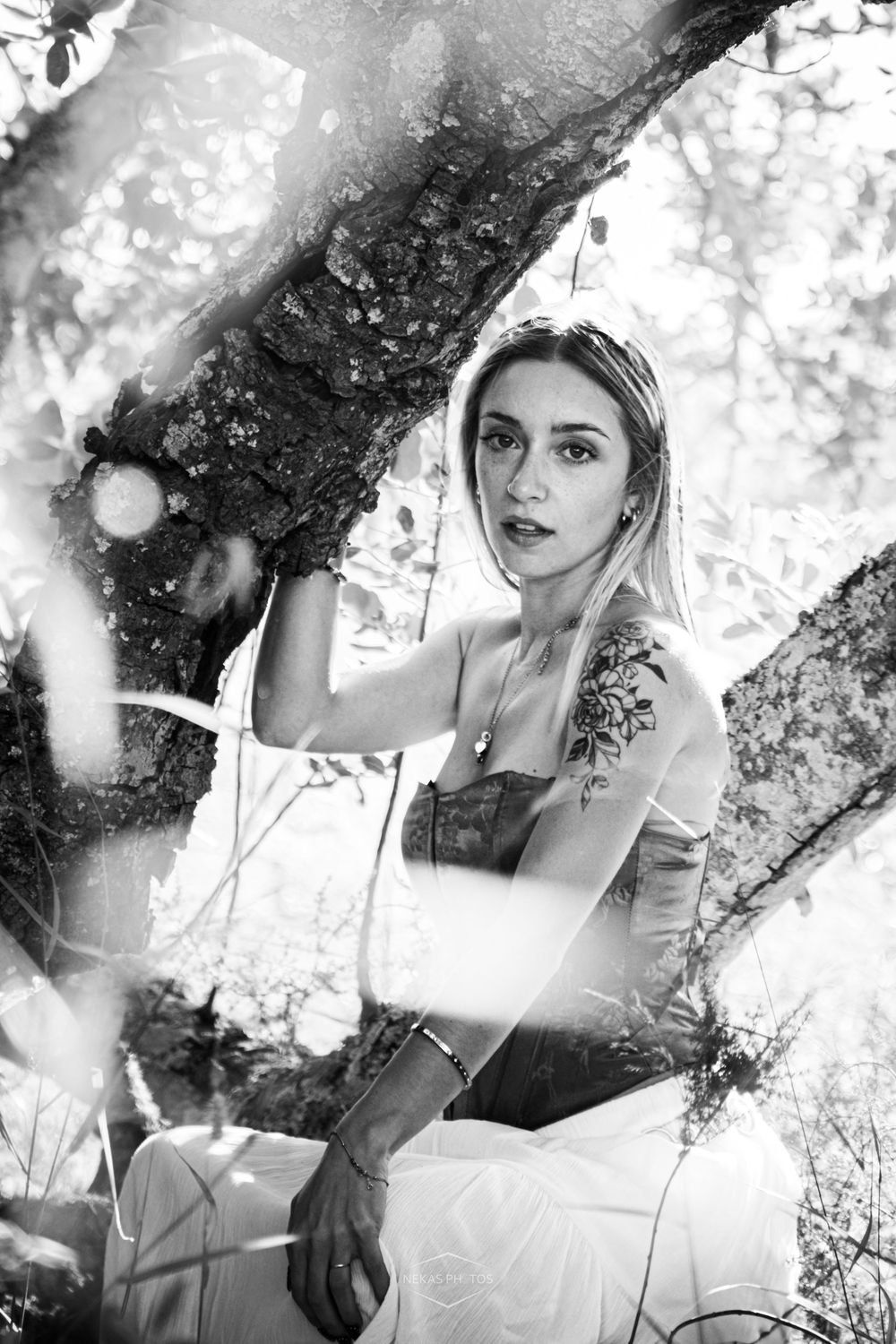 Black and white portrait of a young woman with tattoos sitting among tree branches in a forest setting.