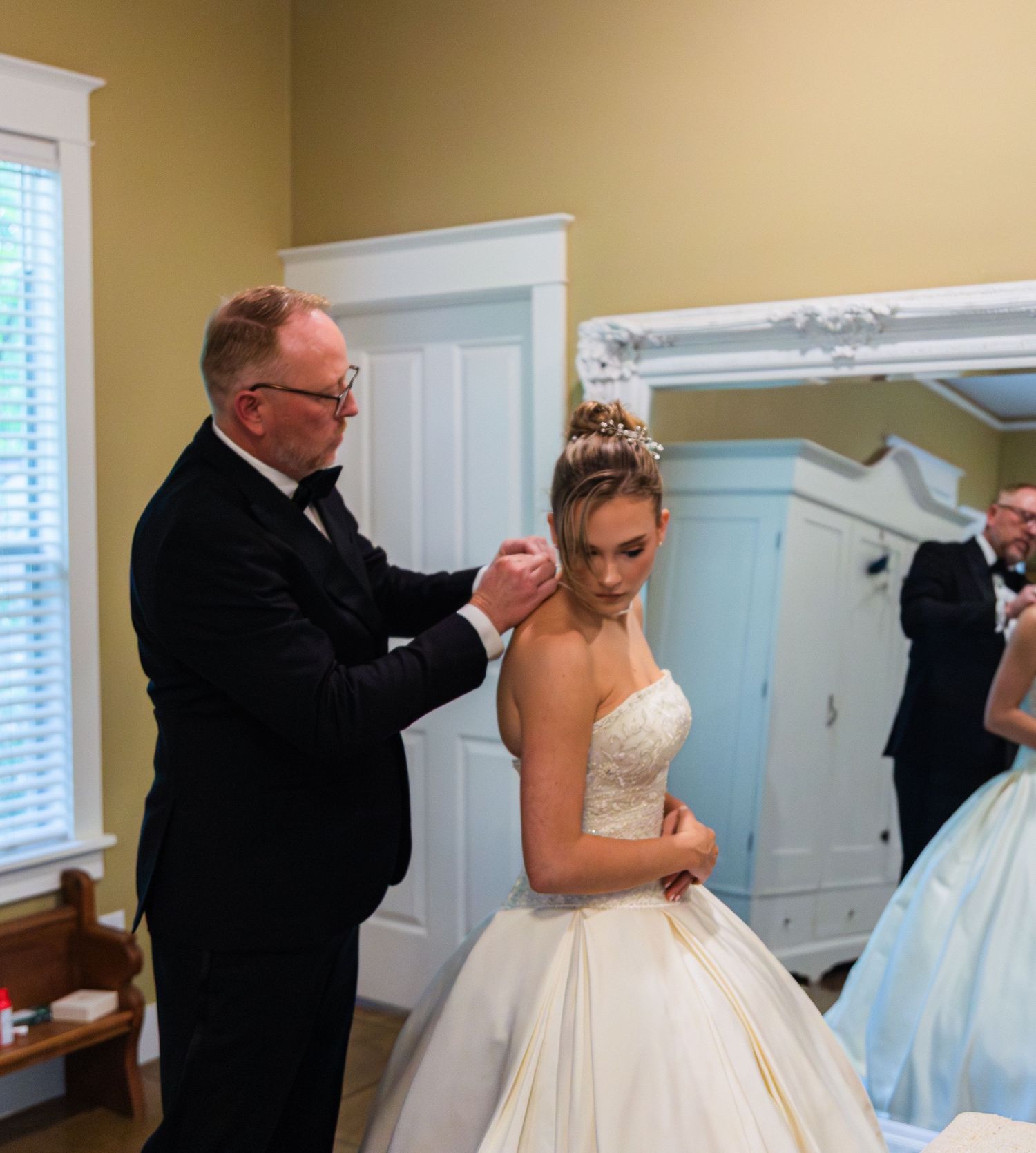 Father helping bride with necklace in white wedding gown before ceremony in elegant room with mirror.
