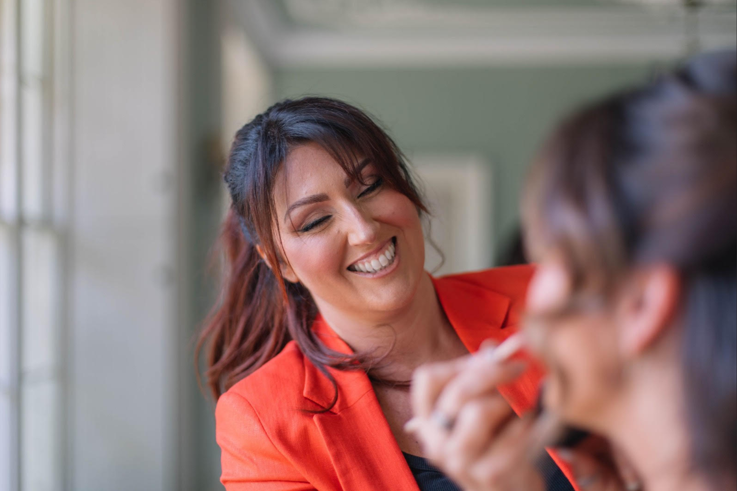 A smiling woman in a red blazer applies makeup to another person in a bright, elegant setting.