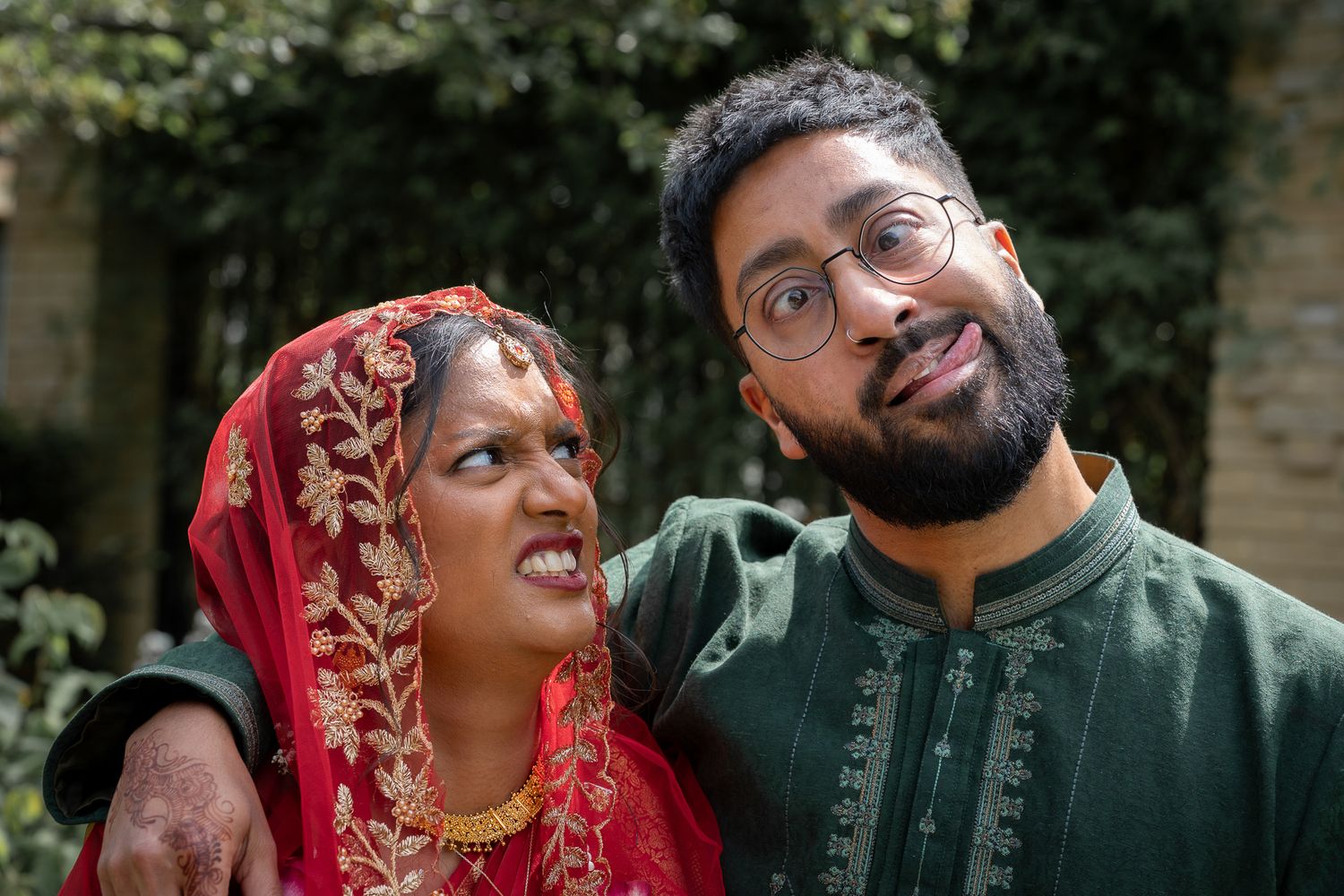 South Asian couple in traditional attire, woman in red bridal saree and man in green kurta, posing outdoors.