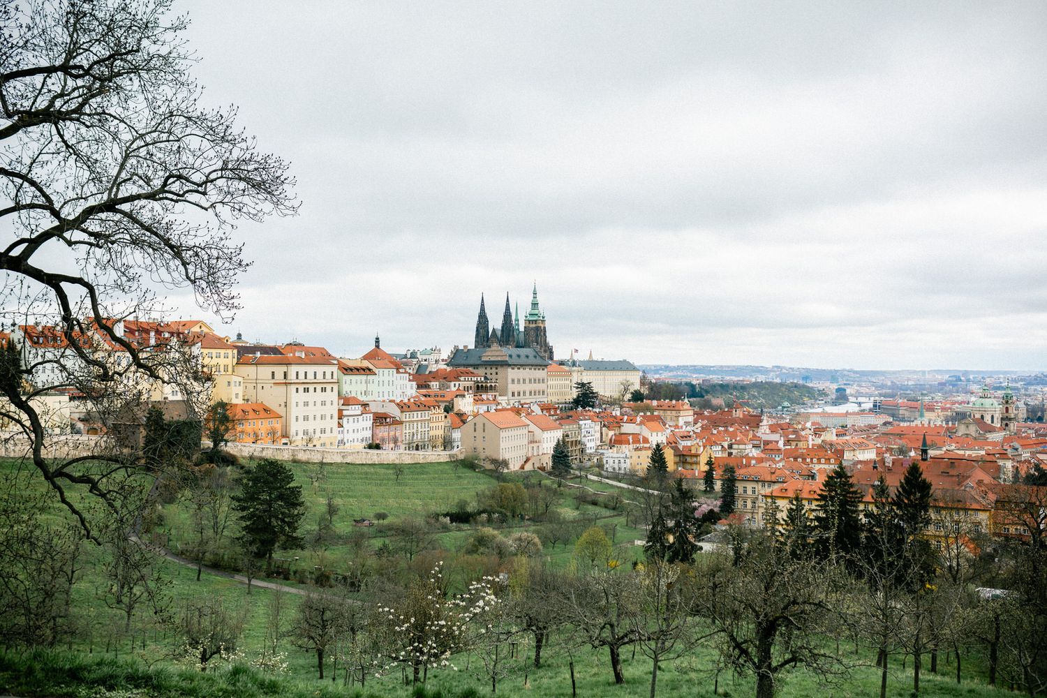 Prague Castle and St. Vitus Cathedral dominate the city skyline on a cloudy day among green hills.