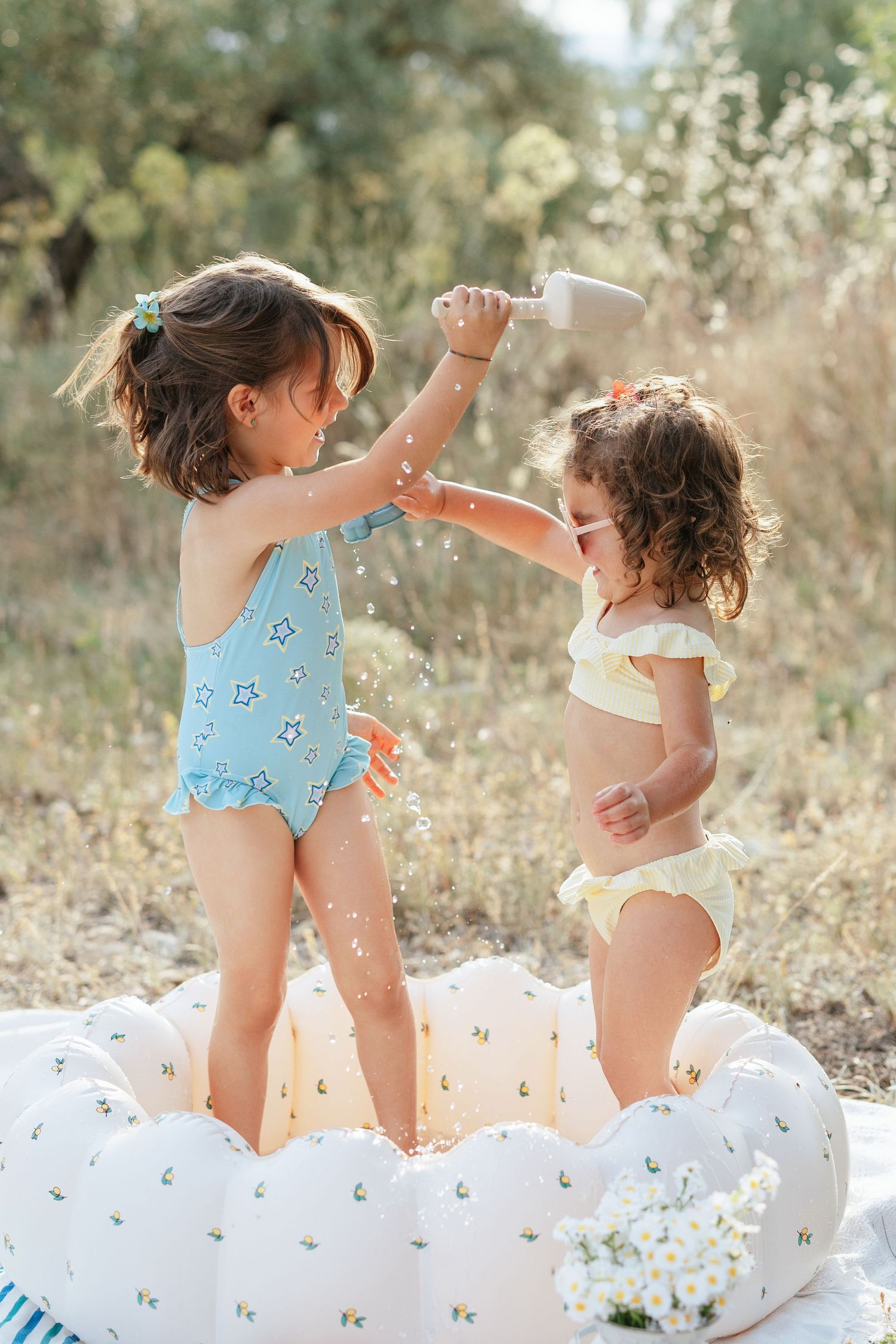 Two young girls in swimsuits playing with water in an inflatable pool outdoors on a sunny day.