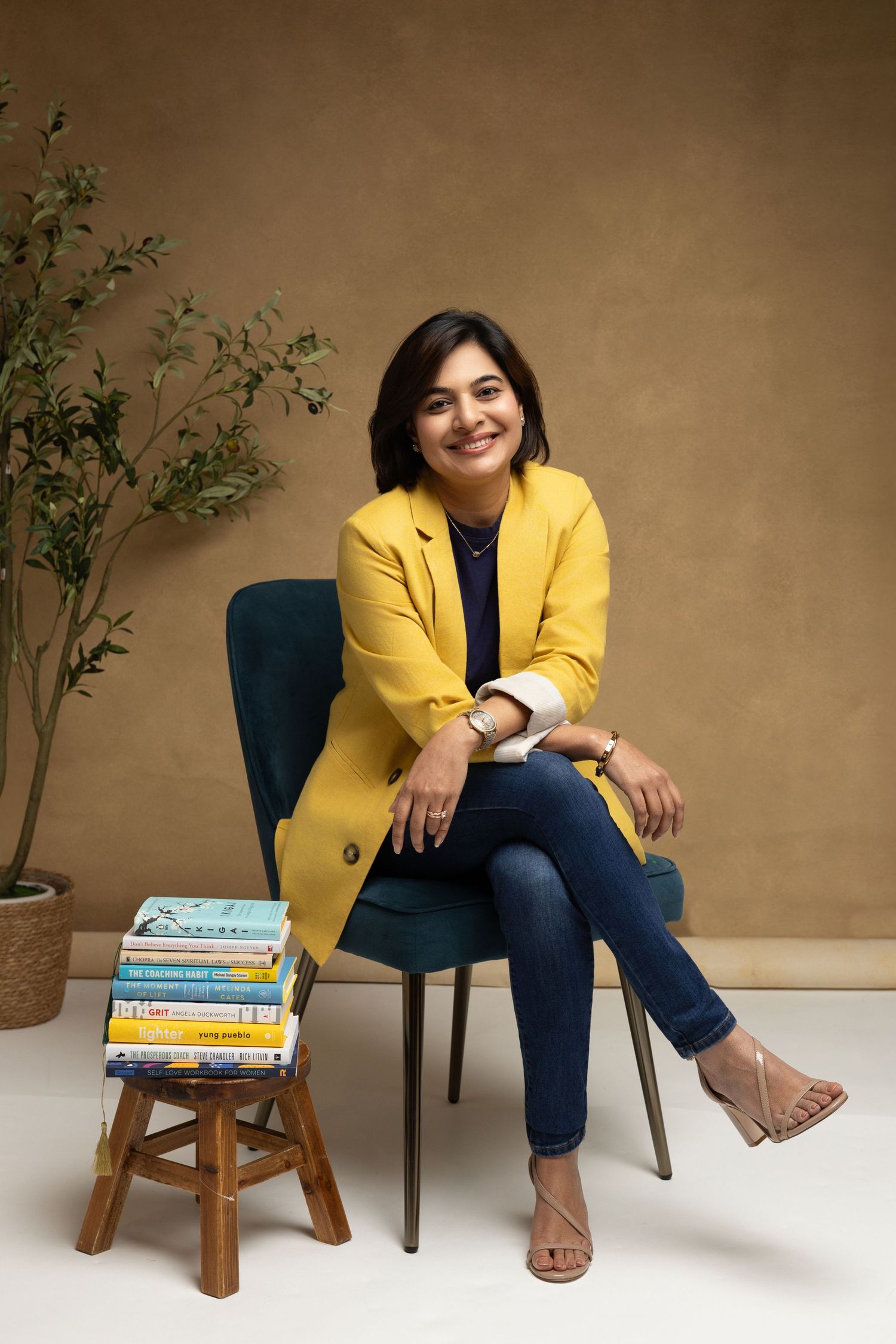 Woman in yellow blazer and jeans sitting on teal chair beside stack of books against tan wall.