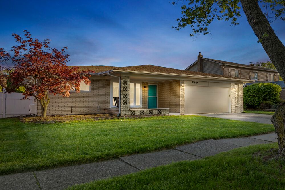 Beige brick ranch home with teal front door, attached garage, red maple tree, and green lawn at dusk.