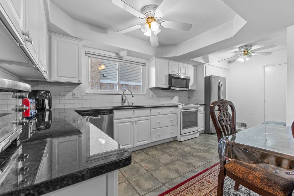 White kitchen with dark granite counters, tile backsplash, stainless appliances, ceiling fan, and tray ceiling.
