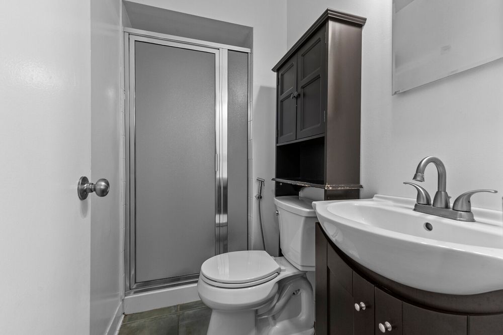 Compact bathroom with glass shower stall, toilet, dark wood vanity, white sink, and overhead storage cabinet.