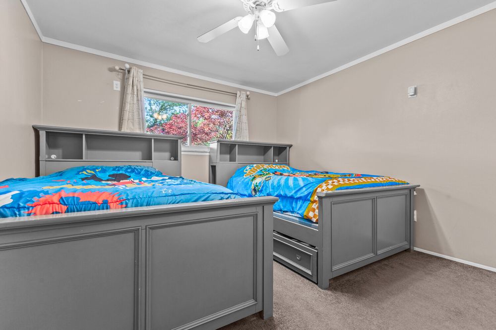 Cozy bedroom with two gray twin beds, built-in shelving headboards, carpet, ceiling fan, and bright window.