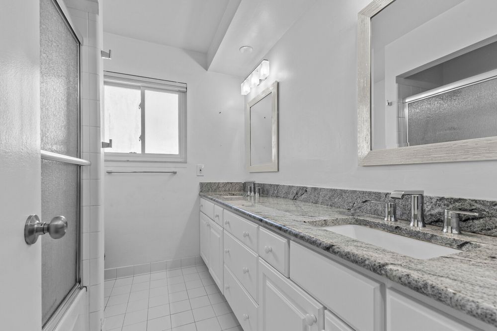 White bathroom with double granite countertop vanity, large mirror, and frosted glass shower door.
