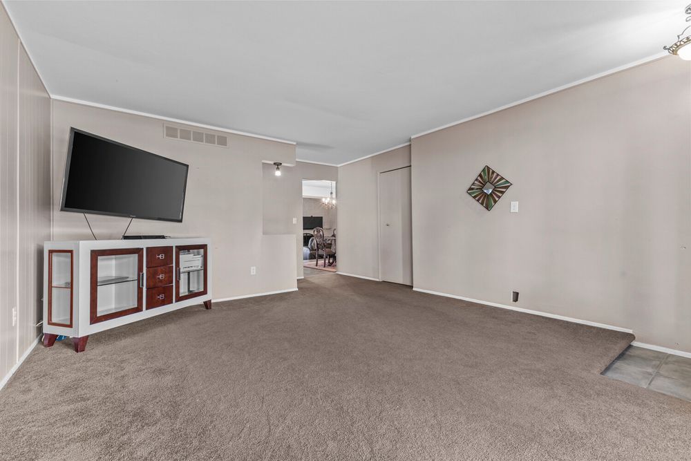 Spacious living room with neutral carpet, wall-mounted flat-screen TV, white trim, and an open doorway to adjacent room.